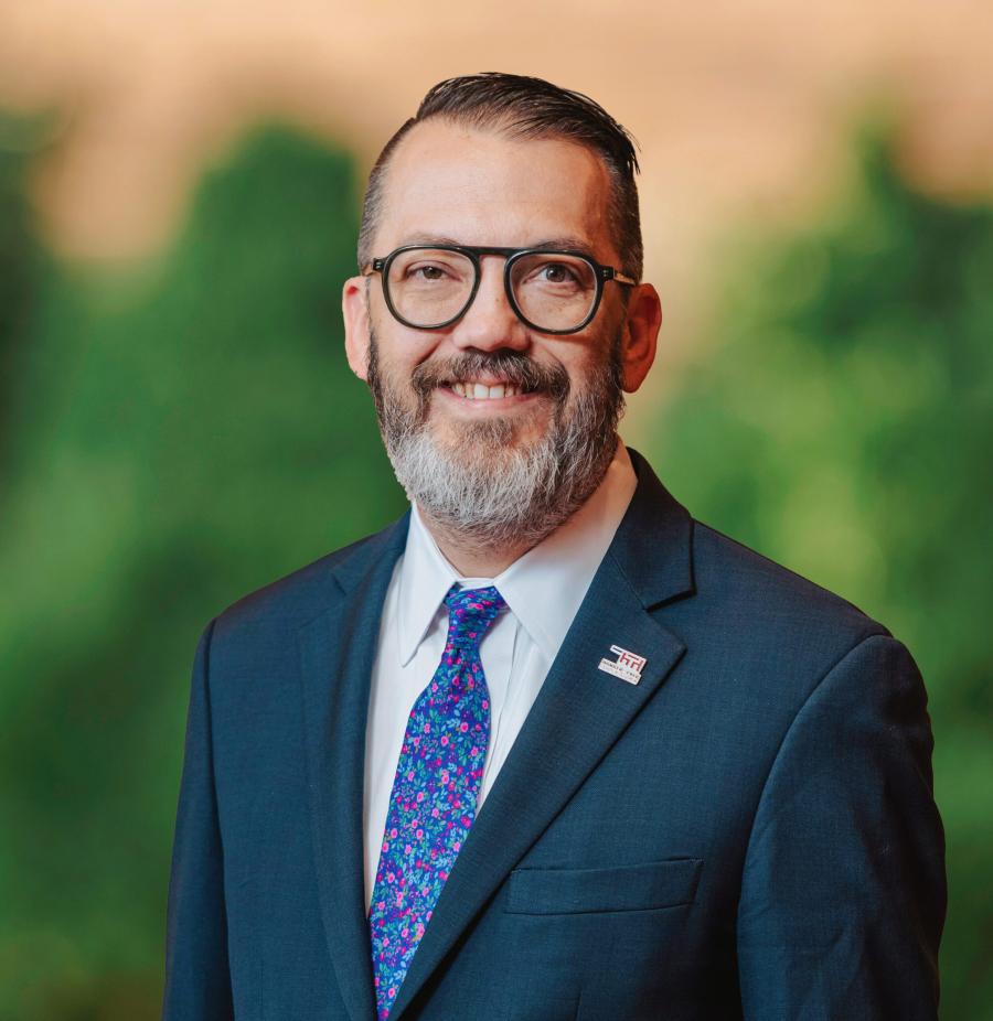 A fair skinned man with black glasses wearing a suit smiling at the camera